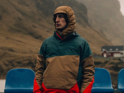 Una persona con una chaqueta verde y marrón está de pie en un paisaje montañoso y nevado con un pequeño edificio de fondo.