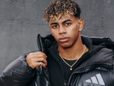 Un hombre joven de pelo rizado y expresión seria, con una chaqueta negra, está de pie frente a un fondo gris.