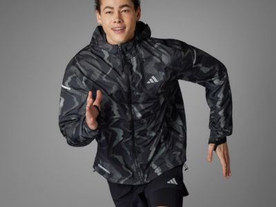 A person wearing a black and gray camouflage-patterned Adidas jacket stands against a plain gray background, gesturing with their hands.