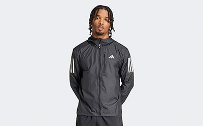 A person wearing a black Adidas jacket with white stripes stands against a plain white background.