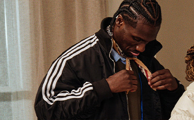 The image shows a person with braided hair wearing a black jacket with white stripes, standing in what appears to be a room with a curtain in the background.