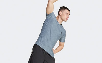 A young man in a gray shirt stands with his arm raised against a plain white background.
