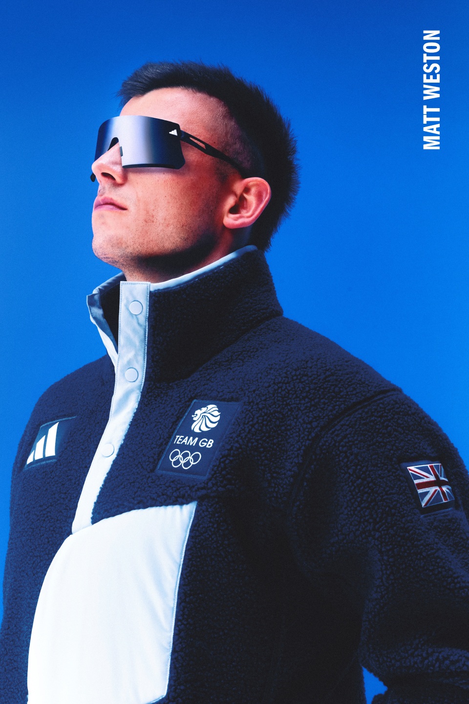 A man wearing a Team GB jacket and sunglasses stands against a vibrant blue sky.