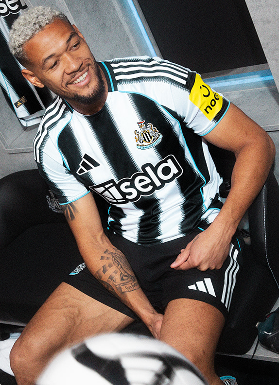 A smiling man wearing a black and white soccer jersey sits in a vehicle, with a microphone in front of him.
