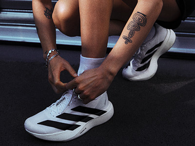 The image shows a person's legs and feet, with the person wearing white sneakers and having a tattoo on their arm.