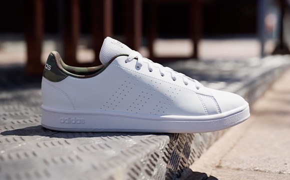 A white Adidas sneaker with green accents sits on a wooden surface, with a blurred background of wooden beams and railings.