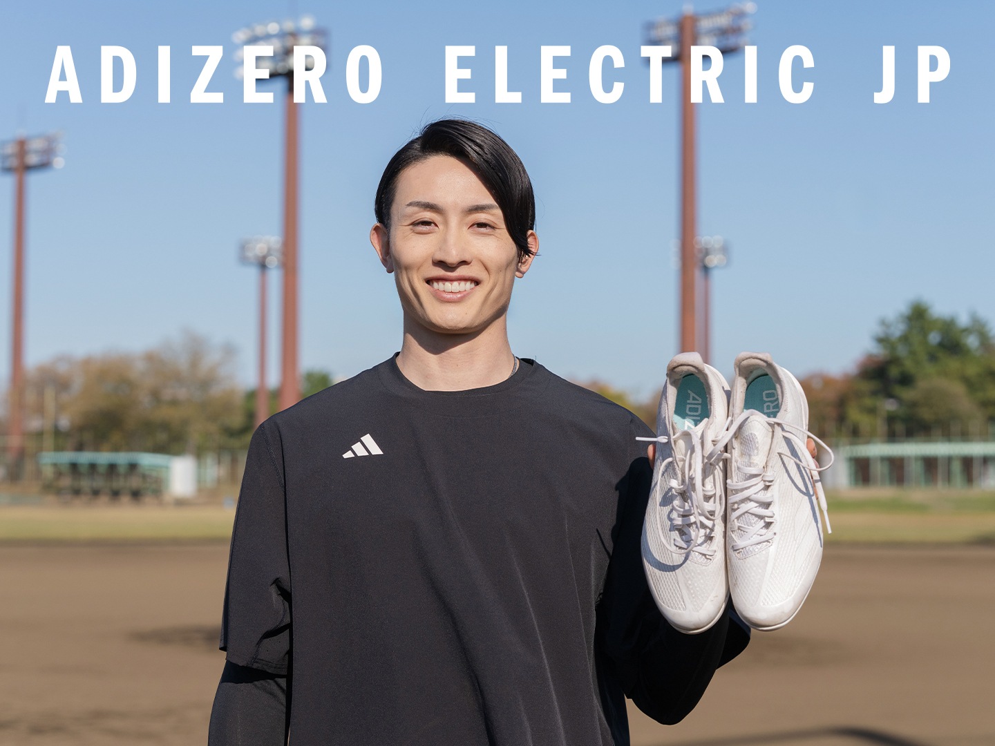 A smiling young man wearing an Adidas shirt and holding a pair of soccer cleats stands on a sports field with stadium lights in the background.