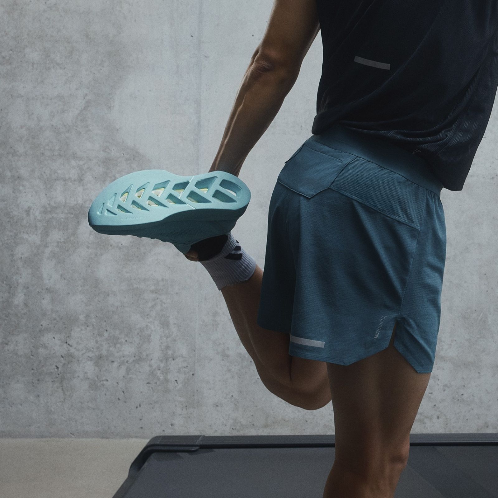 The image shows a person's lower body wearing athletic shorts and shoes with a distinctive teal-colored sole, standing in front of a plain concrete wall.