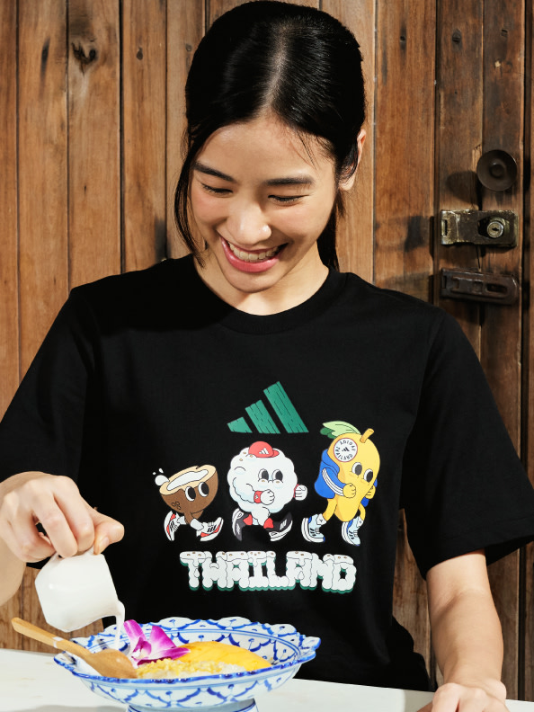 A smiling young woman wearing a black t-shirt with cartoon characters and the word "Thailand" is standing in front of a wooden background.
