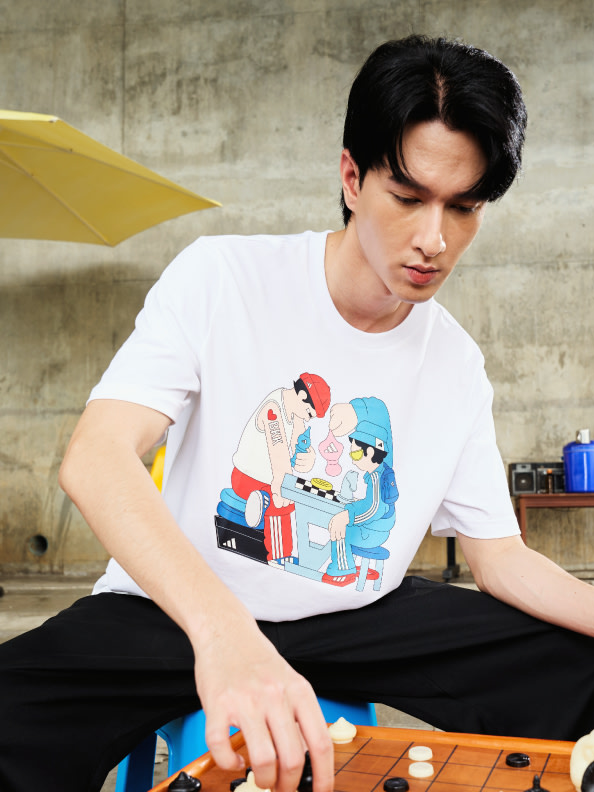 A young man wearing a white t-shirt with a colorful cartoon design sits on a wooden bench in front of a concrete wall.
