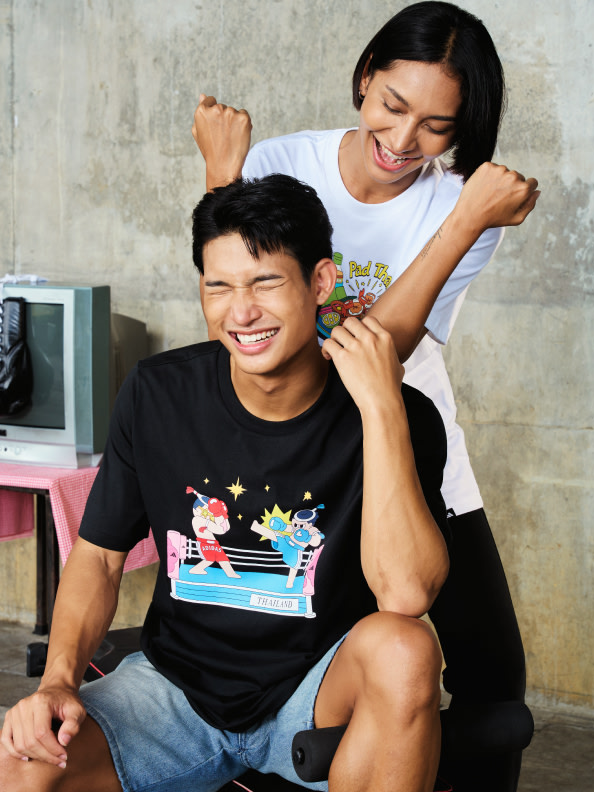A smiling young woman is embracing a man wearing a graphic t-shirt, with a simple background of a concrete wall and some household items visible.