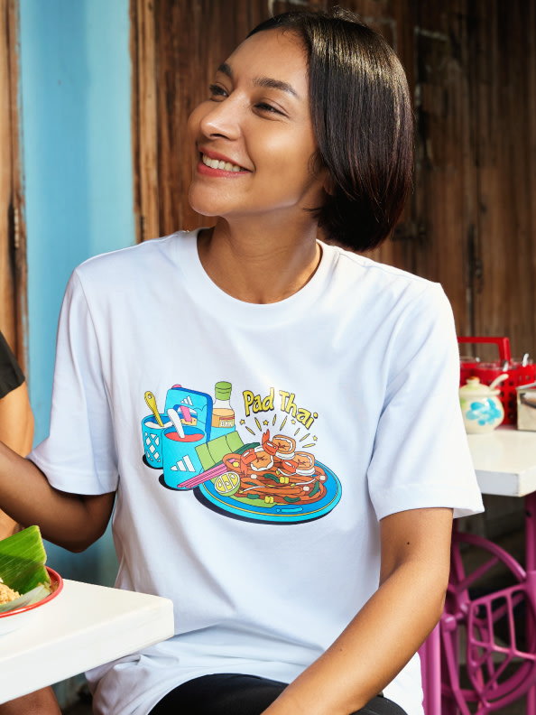 A smiling young woman wearing a white t-shirt with a colorful food-themed graphic, sitting at a table in what appears to be a casual dining setting.