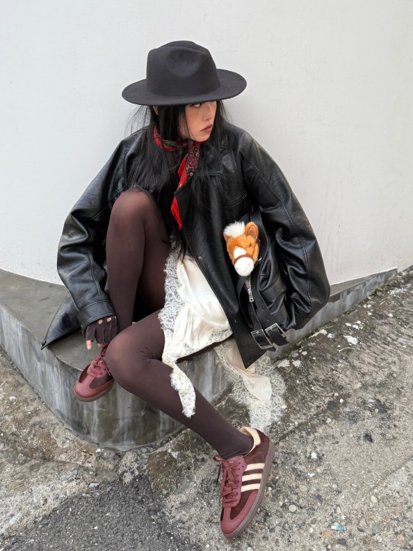 A person wearing a black hat, leather jacket, and red stockings sits on a concrete structure against a plain background.