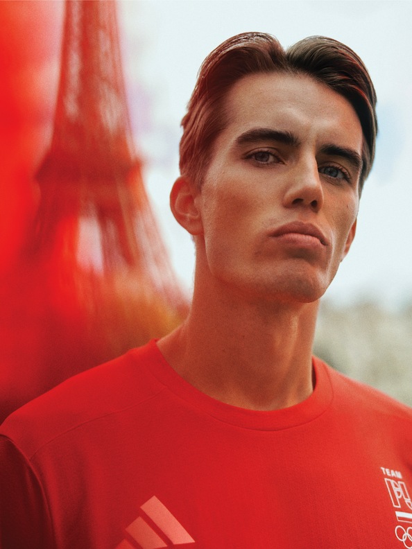 Młody mężczyzna z krótkimi ciemnymi włosami w czerwonej koszulce stoi przed czerwoną konstrukcją, prawdopodobnie Wieżą Eiffla, w outdoorze.