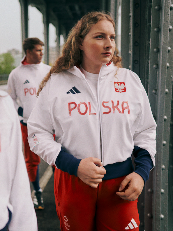 Na zdjęciu młoda kobieta w białej kurtce z nadrukowanym słowem "POLSKA", stojąca przed metalowym ogrodzeniem lub barierą, z inną osobą widoczną w tle.