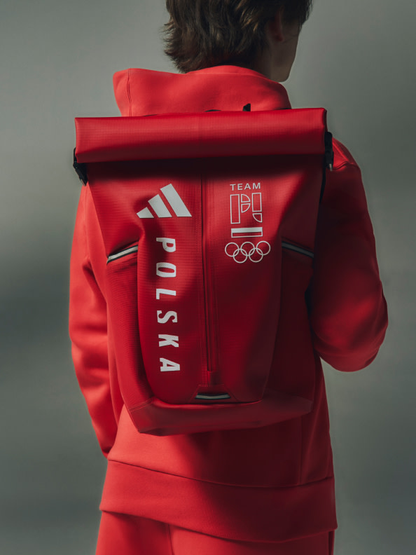 Osoba w czerwonej bluzie reprezentacji Polski z logo olimpijskim, stojąca na szarym tle.