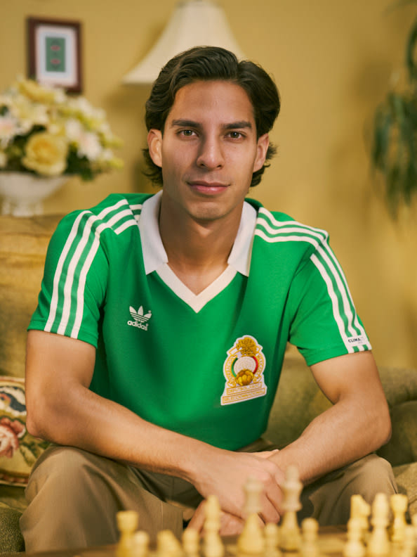 Un hombre joven con una camiseta deportiva verde con un logotipo está sentado en una habitación con arreglos florales y una ilustración enmarcada en las paredes.