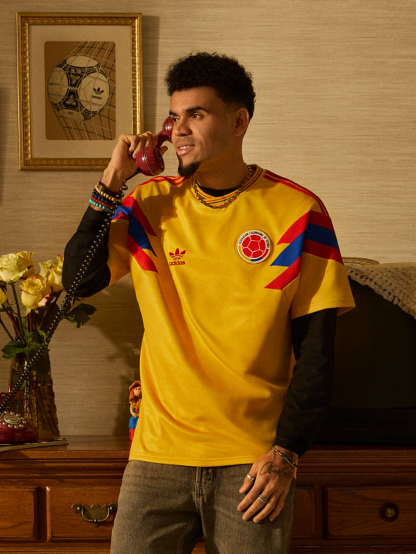 Un hombre joven con una vibrante camiseta deportiva amarilla y roja está de pie en una habitación con una ilustración enmarcada en la pared y varios artículos decorativos en una cajonera de madera detrás de él.