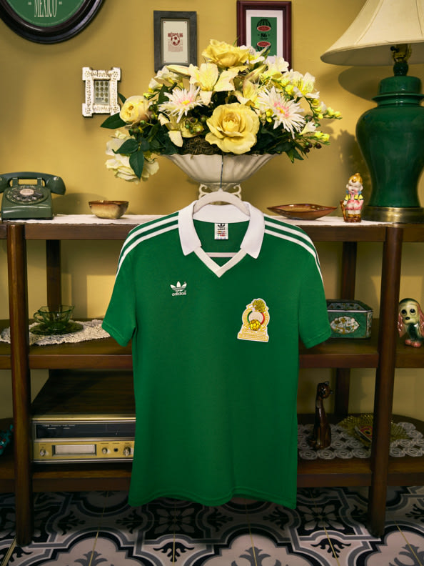 La imagen muestra una camiseta de fútbol verde exhibida en un estante, con un jarrón de flores amarillas y varios artículos decorativos de fondo.