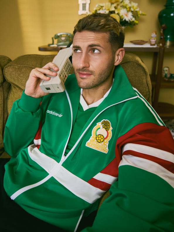 Un hombre con una camiseta deportiva verde está sentado en un sofá y hablando por un teléfono móvil, con varios elementos decorativos visibles en el fondo.