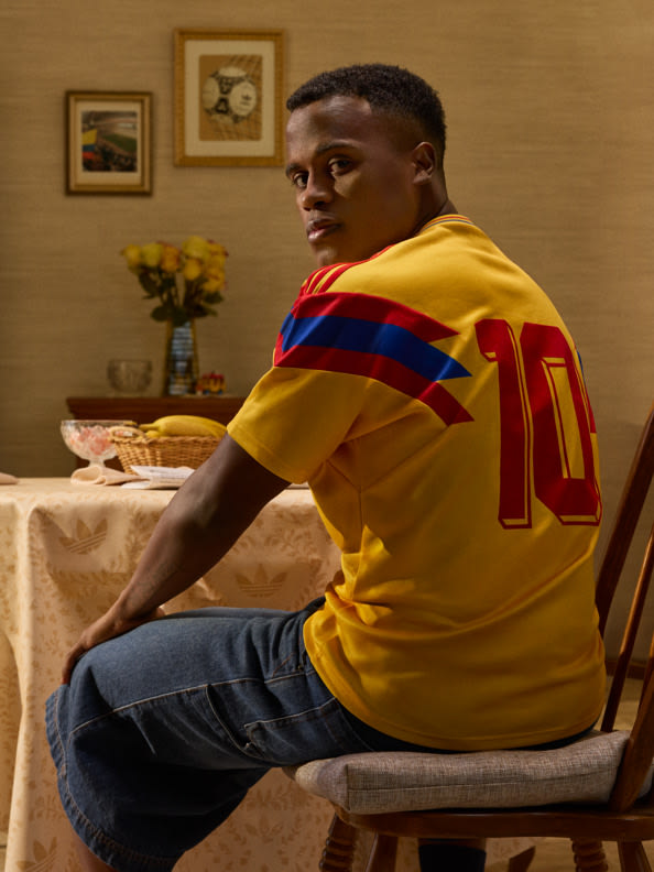 Un joven vestido con una camiseta amarilla con el número 10 está sentado en una mesa en una habitación con una ilustración enmarcada en las paredes y un jarrón de flores.