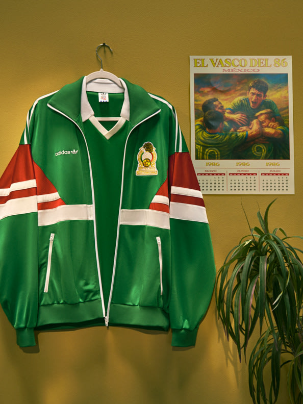 Una chaqueta deportiva verde y blanca cuelga en una pared, con un calendario vintage con una escena vaquera de fondo y una maceta en una esquina.