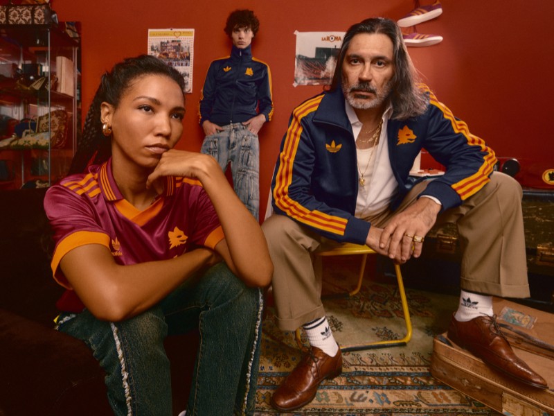 Two individuals, a woman and a man, are seated in a room with vibrant orange walls and various decorative items in the background.