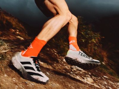 Na zdjęciu widać nogi osoby w jasnopomarańczowych skarpetkach i czarno-białych butach sportowych, biegnącej po zabrudzonym szlaku otoczonym liśćmi.