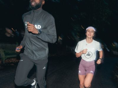 The image shows two people, a tall individual wearing a gray outfit and a shorter individual wearing a white shirt and pink shorts, walking together in a dimly lit outdoor setting.