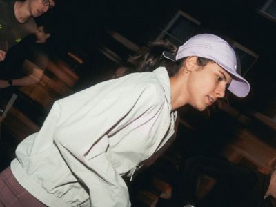 A person wearing a white hooded jacket and cap is standing in a dimly lit environment with what appears to be wooden structures or buildings in the background.
