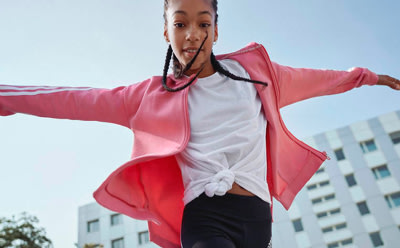 Una persona con una chaqueta rosa brillante se coloca de pie con los brazos estirados sobre un fondo de edificios modernos.