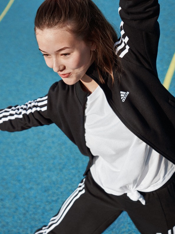 Eine junge Frau in einem schwarz-weißen Sportoutfit steht auf einem blauen Sportplatz und ihre Haare wehen im Wind, während sie ihre Arme ausstreckt.