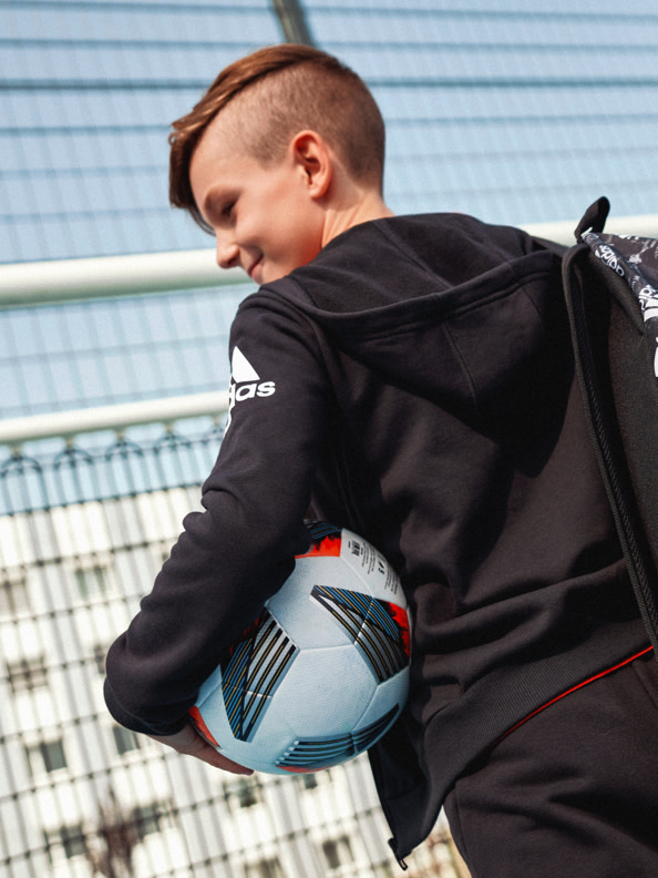 Ein junger Mensch in einer schwarzen Sportjacke hält einen Fußball und steht vor einem eingezäunten Sportplatz.