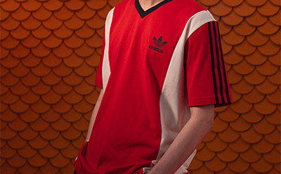 A vibrant red jersey with the Adidas logo stands out against a backdrop of overlapping orange scales, creating a visually striking contrast.