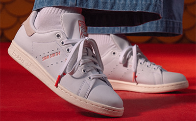 A pair of white sneakers with red accents sits on a red surface, with a denim-clad leg visible in the background.