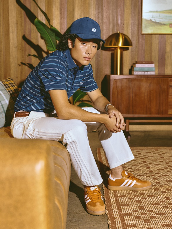 A young man wearing a blue striped shirt, white pants, and orange sneakers is sitting on a couch in a room with wooden paneling and a lamp in the background.