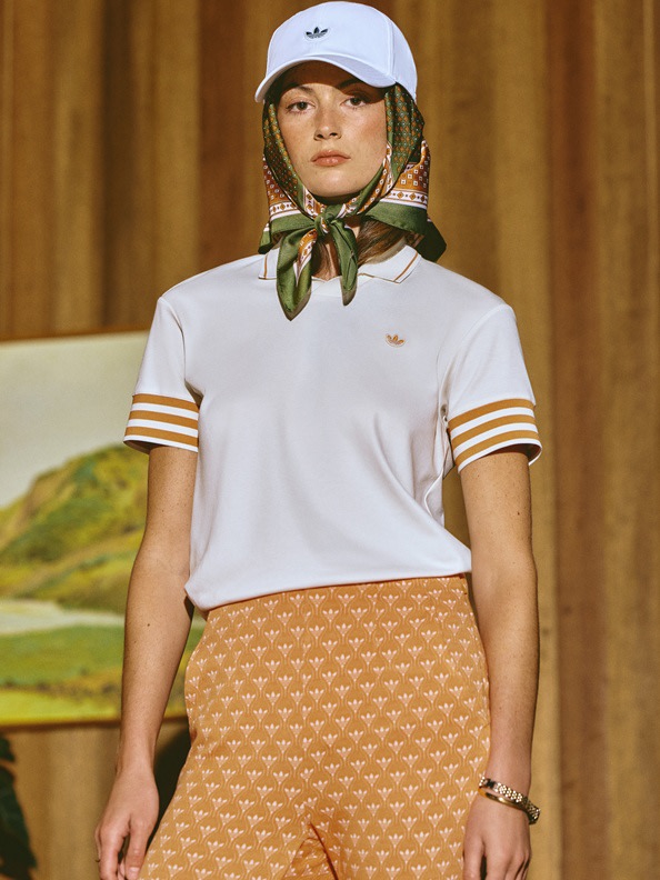 A young woman wearing a white shirt, a patterned scarf, and a baseball cap stands in front of a painted landscape backdrop.