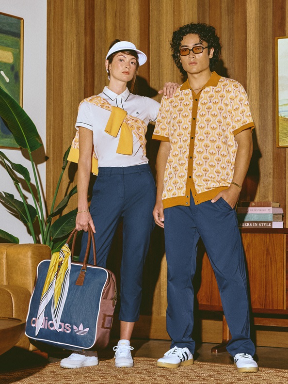 Two individuals, a woman and a man, are standing in a room with wooden paneling on the walls. The woman is wearing a white shirt, navy blue pants, and a white hat, while the man is wearing a patterned yellow shirt and navy blue pants. They both appear to be holding bags, with the woman carrying an Adidas-branded bag.