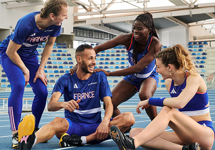 Un groupe d'athlètes en uniforme bleu, hommes et femmes, sont rassemblés sur un terrain de sport indoor avec des sièges bleus et blancs en arrière-plan.