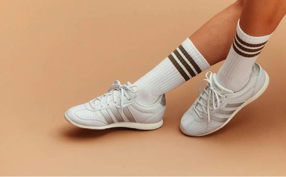 A pair of white sneakers with three black stripes on the side, worn with white socks, against a peach-colored background.