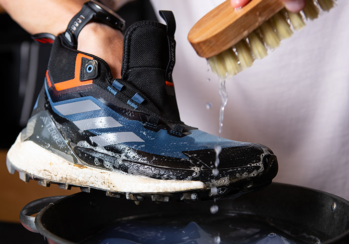 A dirty, worn-out hiking boot is being scrubbed with a stiff-bristled brush, with water splashing onto the surface.