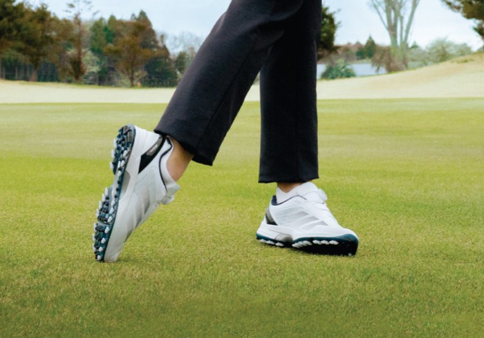 The image shows a person's lower body wearing black pants and white golf shoes standing on a lush green golf course with trees and foliage in the background.