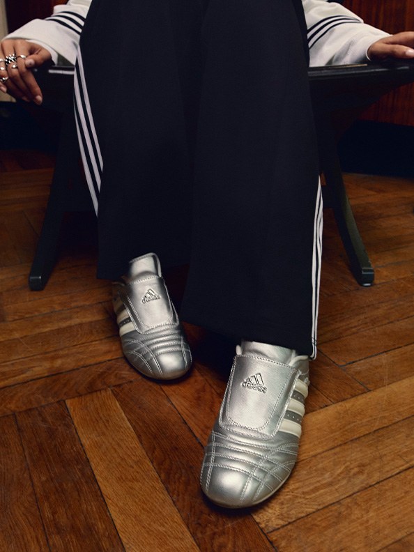 The image shows a person wearing a black suit and silver-colored Adidas shoes, standing on a wooden floor.