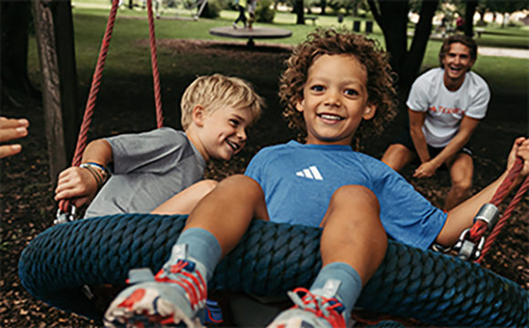 Two young children, a boy and a girl, are happily playing on a swing set in a park-like setting with trees and other playground equipment visible in the background.