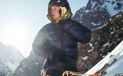 Člověk v tmavé bundě s kapucí stojí v hornaté zasněžené krajině s kamenitými útesy v pozadí.