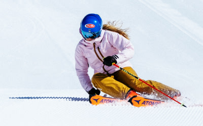 Člověk v růžové bundě, modré helmě a žlutých kalhotách lyžuje na zasněženém svahu s jasnou oblohou na pozadí.