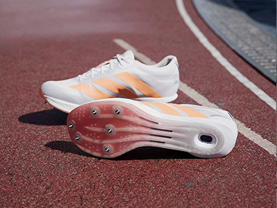 A pair of white and orange athletic shoes with spikes on the soles rests on a red running track.