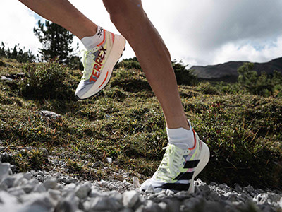 The image shows a person's legs and feet wearing white and colorful athletic shoes standing on a rocky, grassy terrain with mountains in the background.