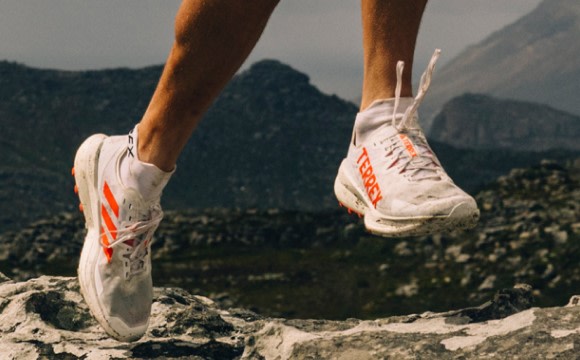 The image shows a pair of athletic shoes with orange and white accents, worn by a person standing on a rocky, mountainous terrain with lush greenery in the background.