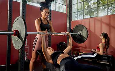 A woman in athletic attire is assisting another person with a weight lifting exercise in a well-equipped gym setting with large windows providing natural light.